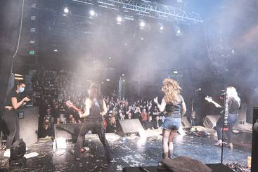 Eine Rockband spielt auf einer Bühne vor einem begeisterten Publikum. Drei Musikerinnen stehen im Vordergrund, während die Zuschauer im Hintergrund den Auftritt genießen. Der Raum ist von Nebel erfüllt, und die Atmosphäre ist lebhaft und energiegeladen.