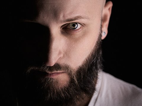 Ein bärtiger Mann mit Glatze schaut direkt in die Kamera. Sein Gesicht ist teilweise im Schatten, was einen dramatischen Effekt erzeugt. Der Mann trägt ein schlichtes, weißes T-Shirt. Die Beleuchtung hebt seine markanten Gesichtszüge hervor.