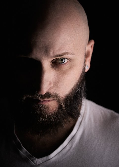 A close-up portrait of a bald man with a thick beard, staring intensely at the camera. The lighting creates a dramatic effect, with one side of his face in shadow and the other illuminated, highlighting his facial features and expression. He wears a simple white shirt.