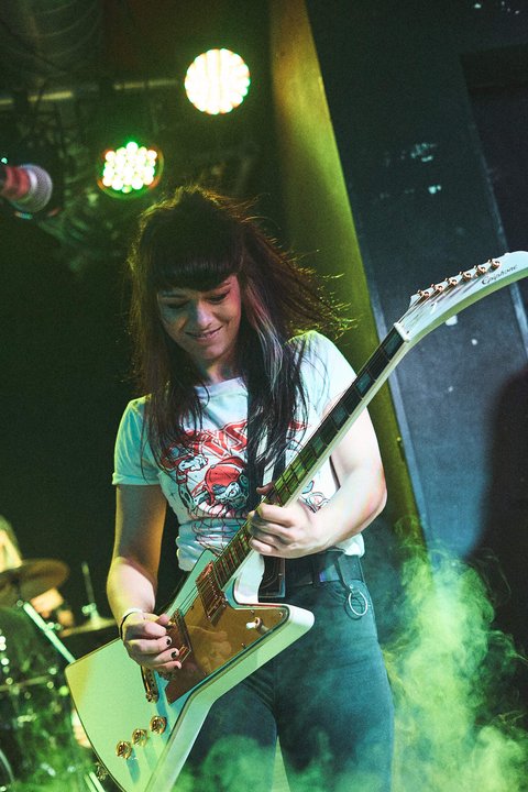 Eine junge Frau spielt leidenschaftlich eine elektrische Gitarre auf der Bühne. Sie trägt ein T-Shirt mit einem auffälligen Aufdruck und hat lange, dunkelhaarige Frisur. Der Hintergrund ist schwach beleuchtet und mit Nebel gefüllt, was eine energiegeladene Atmosphäre schafft.