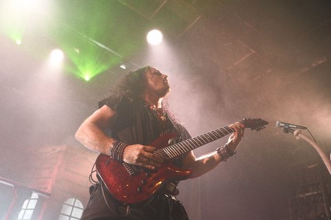 Ein Musiker spielt leidenschaftlich Gitarre auf der Bühne. Er hat langes, dunkles Haar und trägt ein schlichtes schwarzes Outfit. Scheinwerferlicht beleuchtet ihn, während er sich in die Musik vertieft. Die Atmosphäre ist dynamisch und energisch.