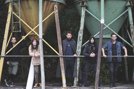 Fünf Männer stehen nebeneinander vor einer rustikalen Kulisse mit großen, bunten silos. Die Gruppe hat verschiedene Stile, von lässig bis mystisch. Die Atmosphäre wirkt geheimnisvoll und urban.