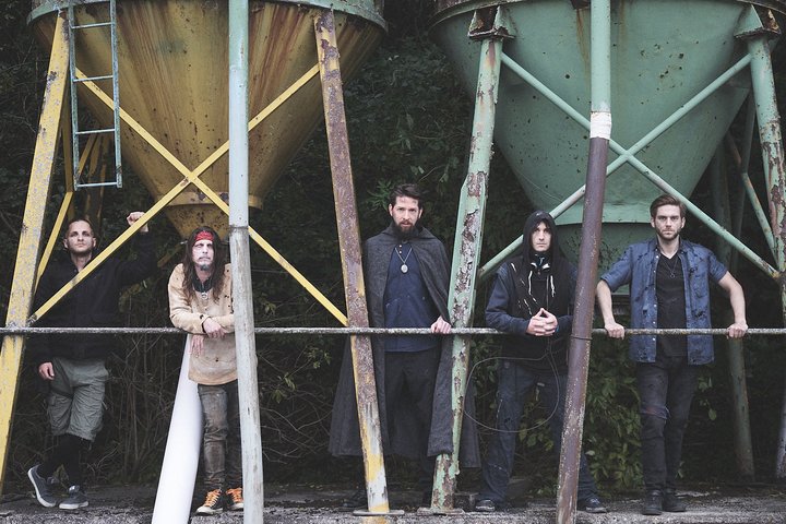 Aeons of Ashes Fünf Männer stehen nebeneinander vor einer rustikalen Kulisse mit großen, bunten silos. Die Gruppe hat verschiedene Stile, von lässig bis mystisch. Die Atmosphäre wirkt geheimnisvoll und urban.