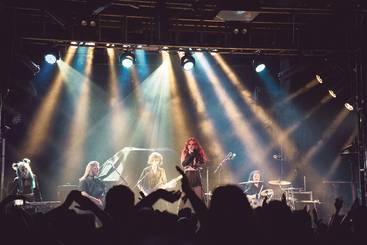 Eine Band steht auf der Bühne, während sie live spielt. Die Sängerin mit rotem Haar steht im Vordergrund, umgeben von anderen Musikern. Scheinwerfer strahlen hell auf die Bühne, während die Menge im Vordergrund ihre Hände hebt und den Auftritt genießt.