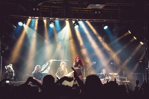 Eine Band steht auf der Bühne, während sie live spielt. Die Sängerin mit rotem Haar steht im Vordergrund, umgeben von anderen Musikern. Scheinwerfer strahlen hell auf die Bühne, während die Menge im Vordergrund ihre Hände hebt und den Auftritt genießt.