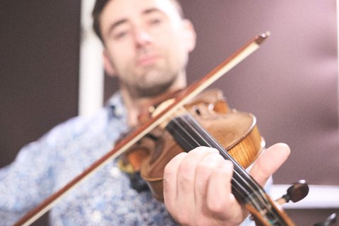 Ein Mann spielt Geige. Er hält den Bogen in der rechten Hand und die Geige in der linken Hand. Der Hintergrund ist unscharf, der Fokus liegt auf dem Musiker und seinem Instrument. Der Mann trägt ein gemustertes Hemd.