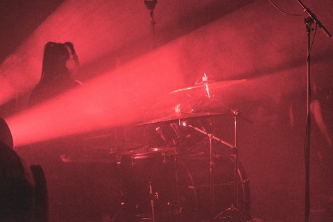 Auf der Bühne ist ein Schlagzeug zu sehen, umgeben von roten Lichtern und Nebel. Eine Person sitzt hinter dem Schlagzeug, während Lichtstrahlen den Raum durchdringen und eine dynamische Atmosphäre schaffen.