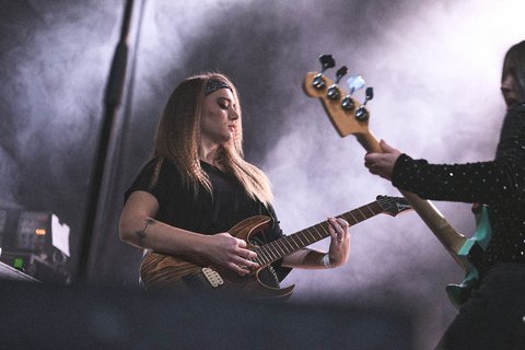 Eine Musikerin spielt leidenschaftlich E-Gitarre auf einer Bühne, umgeben von Nebel. Sie hat lange, glatte Haare und trägt ein schwarzes Oberteil. Neben ihr steht eine weitere Musikerin, die einen Bass spielt. Die Atmosphäre ist energiegeladen und konzentriert.