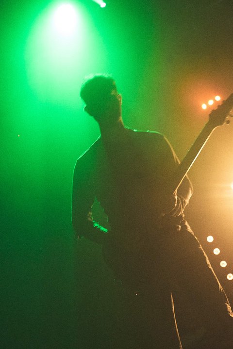 Ein Silhouette eines Musikers, der E-Gitarre spielt, vor bunten Lichtern. Der Künstler ist von grünem und orangefarbenem Licht umgeben, das eine dynamische Bühnenatmosphäre schafft.