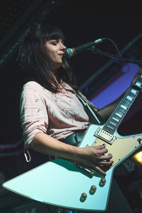 Eine Musikerin mit langen, dunklen Haaren spielt eine elektrische Gitarre auf einer Bühne. Sie trägt ein helles, gemustertes Hemd und singt in ein Mikrofon, während die Bühne schwach beleuchtet ist. Die Stimmung der Szene ist dynamisch und musikalisch.