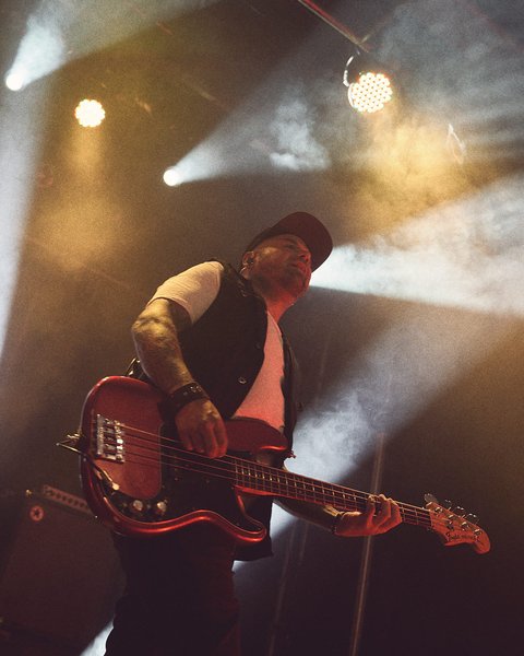 Ein Musiker steht auf einer Bühne und spielt Bass. Er trägt ein schwarzes Outfit und eine Mütze. Scheinwerfer strahlen Licht auf ihn, während er in die Musik vertieft ist.