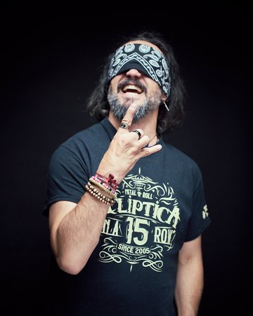 Ein Mann mit einem Bandana, das seine Augen bedeckt, macht die Handzeichen für Rockmusik. Er trägt ein schwarzes T-Shirt mit dem Aufdruck einer Musikgruppe und hat mehrere Armbänder an seinem Handgelenk. Der Hintergrund ist dunkel und hebt seine Pose hervor.