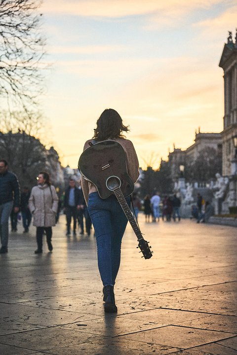 Eine Person mit dunklen Haaren trägt eine Gitarre auf dem Rücken und geht auf einem belebten Platz bei Sonnenuntergang. Im Hintergrund sind Passanten und historische Gebäude sichtbar.