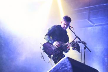 Ein Musiker steht auf der Bühne und spielt E-Gitarre. Er trägt ein auffälliges, schwarzes Outfit und ist von buntem Licht umgeben. Seine Konzentration ist auf die Musik gerichtet, während er inmitten einer Live-Performance ist.