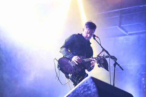 Ein Musiker steht auf der Bühne und spielt E-Gitarre. Er trägt ein auffälliges, schwarzes Outfit und ist von buntem Licht umgeben. Seine Konzentration ist auf die Musik gerichtet, während er inmitten einer Live-Performance ist.