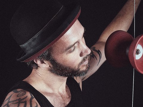 Ein Mann mit Bart und Hut spielt mit einem rotierenden Yo-Yo. Sein Tattoo ist sichtbar, während er konzentriert auf das Spielzeug schaut. Der Hintergrund ist dunkel, wodurch der Fokus auf ihn und das Yo-Yo gelegt wird.