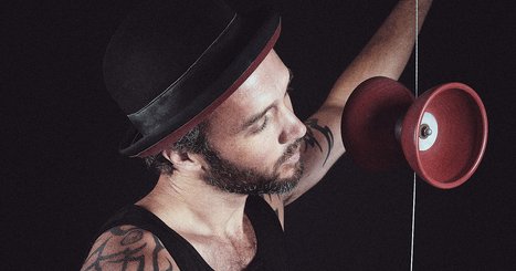 Ein Mann mit Bart und Hut spielt mit einem rotierenden Yo-Yo. Sein Tattoo ist sichtbar, während er konzentriert auf das Spielzeug schaut. Der Hintergrund ist dunkel, wodurch der Fokus auf ihn und das Yo-Yo gelegt wird.