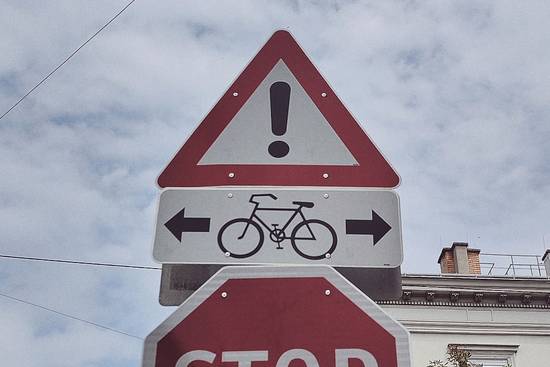 Ein rotes, dreieckiges Verkehrsschild mit einem Ausrufungszeichen oben und einem Fahrradsymbol in der Mitte. Unter dem Fahrradsymbol zeigen zwei Pfeile nach links und rechts, die auf Radfahrer hinweisen. Darunter befindet sich ein Stoppschild.