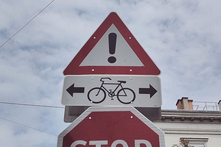 Ein rotes, dreieckiges Verkehrsschild mit einem Ausrufungszeichen oben und einem Fahrradsymbol in der Mitte. Unter dem Fahrradsymbol zeigen zwei Pfeile nach links und rechts, die auf Radfahrer hinweisen. Darunter befindet sich ein Stoppschild.