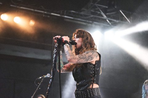 Eine tätowierte Sängerin steht auf der Bühne und hält ein Mikrofon. Sie trägt ein schwarzes Oberteil mit Gürtel und hat lange, lockige Haare. Im Hintergrund sind Lichtstrahlen zu sehen, die die Atmosphäre des Live-Auftritts unterstreichen.