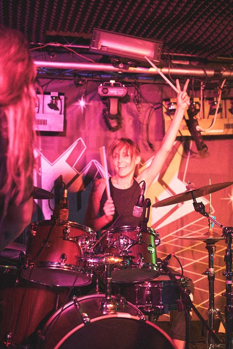 Ein Mädchen spielt begeistert Schlagzeug auf einer Bühne, während sie mit einer Hand das Zeichen für Frieden macht. Die Bühnenbeleuchtung ist farbig und sorgt für eine lebendige Stimmung.