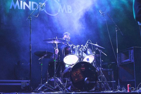 Ein Schlagzeuger spielt leidenschaftlich auf seiner Drumset auf der Bühne, umgeben von Nebel und farbigem Licht. Mikrofone sind aufgestellt, und die Atmosphäre vermittelt die Energie eines Live-Konzerts.