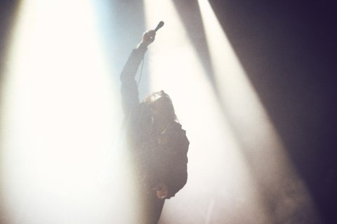 Ein Silhouettenbild eines Sängers, der mit einem Mikrofon in der Hand auf einer Bühne steht. Starke Bühnenlichter strahlen von oben herab und schaffen eine dramatische Stimmung. Die Position des Sängers und die Lichteffekte vermitteln Energie und Intensität eines Live-Auftritts.