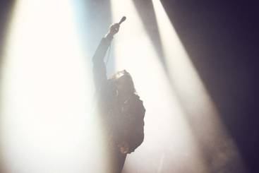 Ein Silhouettenbild eines Sängers, der mit einem Mikrofon in der Hand auf einer Bühne steht. Starke Bühnenlichter strahlen von oben herab und schaffen eine dramatische Stimmung. Die Position des Sängers und die Lichteffekte vermitteln Energie und Intensität eines Live-Auftritts.