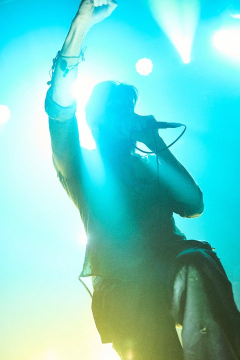 Ein Sänger steht auf der Bühne, während er mit erhobener Hand und Mikrofon in der anderen hand energetisch eine Performance gibt. Die Szene ist von blauen und grünen Lichtern durchzogen, was eine lebhafte Atmosphäre schafft.