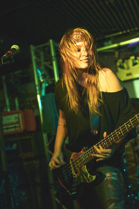 Eine Musikerin spielt leidenschaftlich Bass auf einer Bühne. Ihr längeres Haar weht wild im Rhythmus der Musik, während sie sich in die Darbietung vertieft. Die Beleuchtung ist schwach, mit einem Fokus auf ihrer energiegeladenen Präsenz.