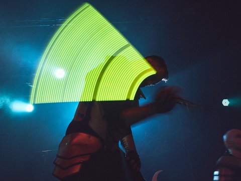 Eine silhouettenhafte Figur steht auf der Bühne, während sie ein Mikrofon hält. Im Hintergrund leuchtet ein grünes, gebogenes Licht, das eine dynamische Atmosphäre schafft. Die Szene vermittelt das Gefühl von Energie und Bewegung während einer Live-Performance.