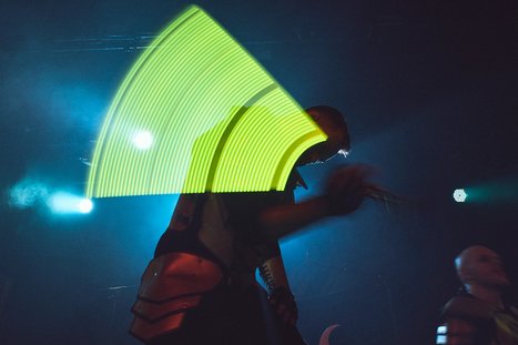 A performer is on stage, holding an illuminated object that creates a vibrant green arc in the air. The background is dark, with blue lighting enhancing the energetic atmosphere of the concert. The focus is on the motion and visual impact of the light.